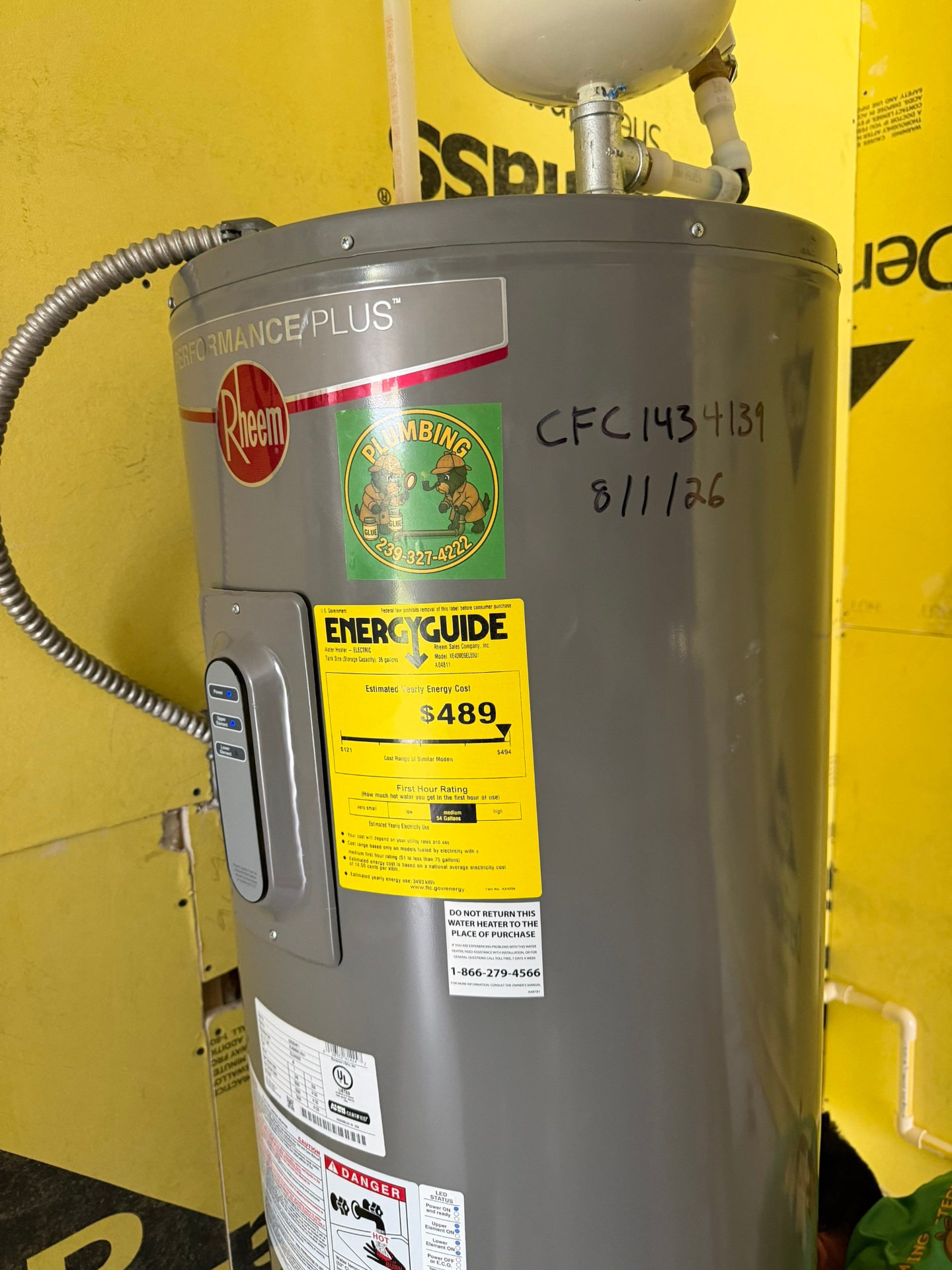 Rheem Performance Plus electric water heater with expansion tank overhead and the license number and install date written on the jacket